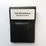 Skinnmappe med navneskilt for sykepleier. Den klassiske skiltmappen brukt av sykepleiere og helsepersonell i mange år. Med skinnlomme for penner, saks osv. Festes i brystlomme på uniform.